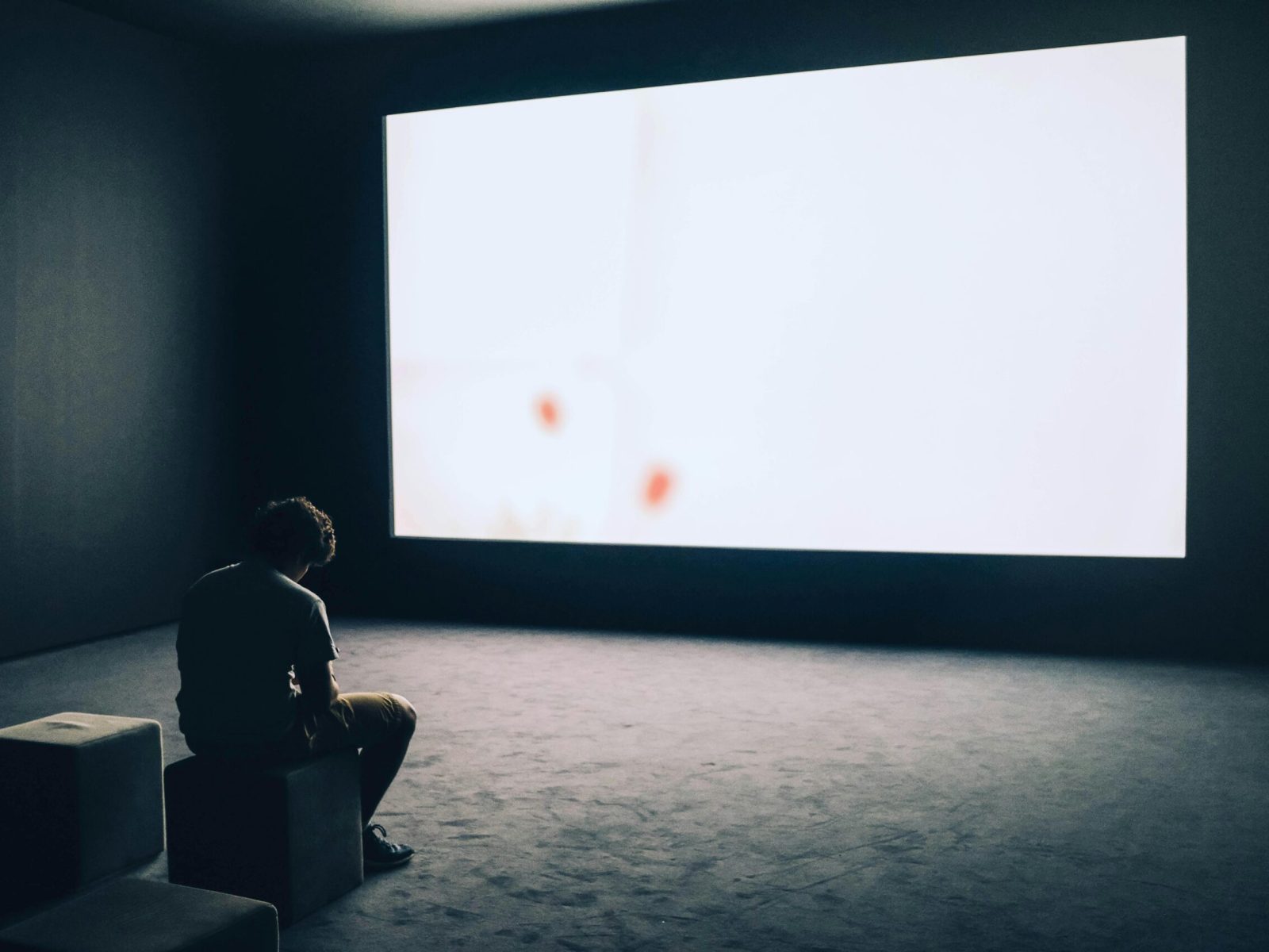 A man sits alone in a dark room with a large white screen, depicting solitude and contemplation.