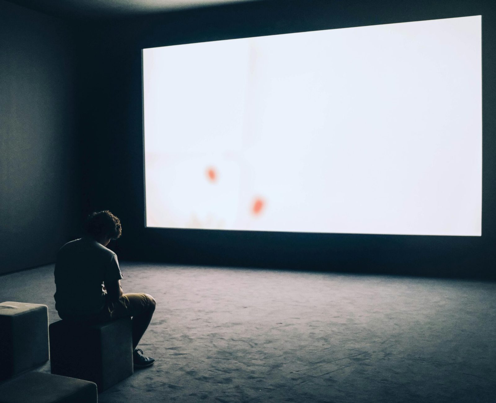 A man sits alone in a dark room with a large white screen, depicting solitude and contemplation.